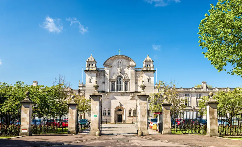 Historisches weißes Kirchengebäude mit zwei Türmen hinter einem Eisenzaun, unter blauem Himmel und umgeben von grünen Bäumen.