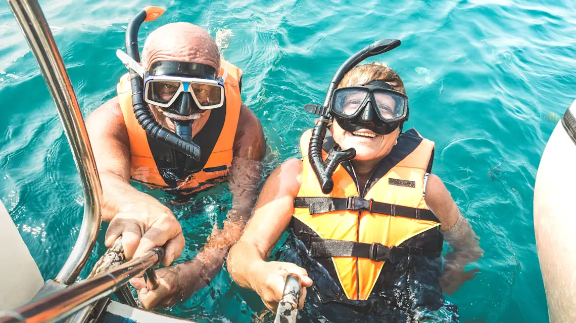 Zwei Schnorchler mit Schwimmwesten und Taucherbrillen im türkisblauen Meerwasser, die sich an einer Bootsleiter festhalten.