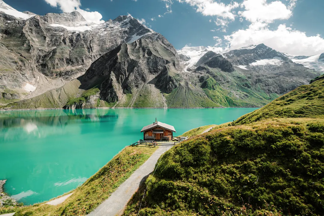 Holzhütte am türkisfarbenen Bergsee, umgeben von majestätischen schneebedeckten Alpen unter blauem Himmel mit weißen Wolken.