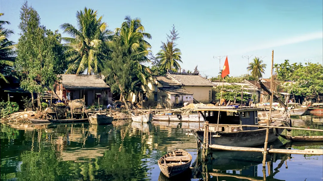 Holzboote auf einem Fluss mit traditionellen Häusern und Palmen im Hintergrund. Mekong-Delta, Vietnam.