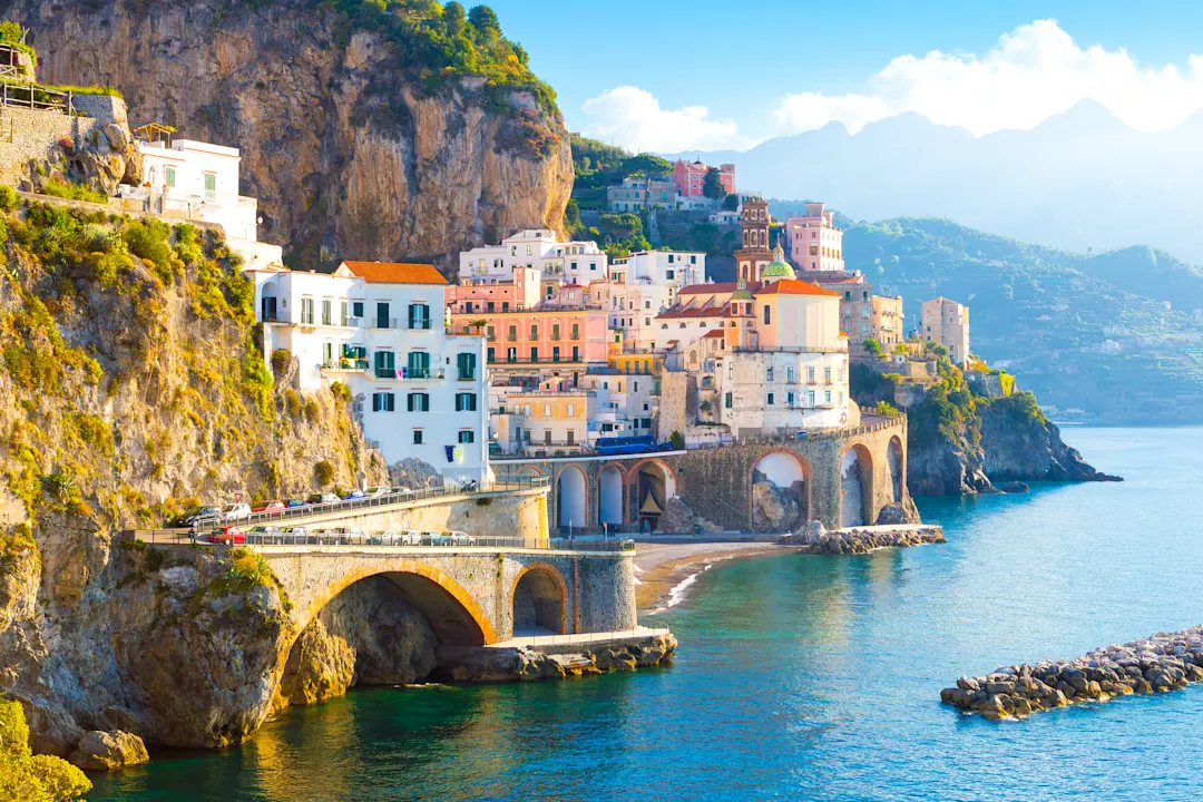 Malerisches Küstendorf Amalfi mit bunten Häusern an steilen Klippen, Steinbrücken über türkisblauem Meer und Bergen im Hintergrund.