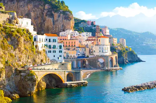 Malerisches Küstendorf Amalfi mit bunten Häusern an steilen Klippen, Steinbrücken über türkisblauem Meer und Bergen im Hintergrund.