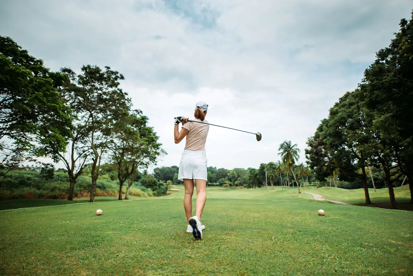 Golf, Angleterre Golfeuse en jupe blanche et haut beige effectuant un swing sur un parcours verdoyant bordé d'arbres sous un ciel nuageux.