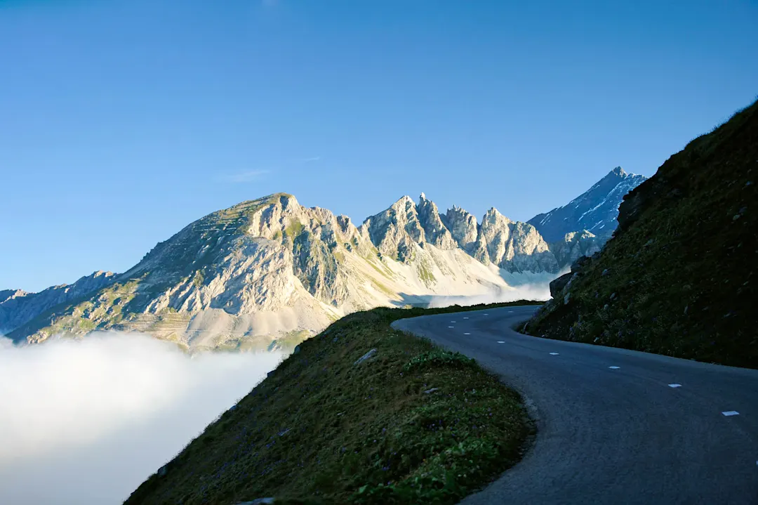 Kurvenreiche Bergstraße mit Blick auf majestätische Alpengipfel über einer Nebelschicht unter klarem blauem Himmel.