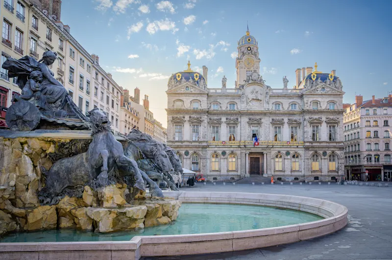 Bartholdi Fountain, Lyon Historisches Rathaus von Lyon mit prachtvoller Fassade und Turmuhr, davor ein Brunnen mit Skulpturen unter blauem Himmel.
