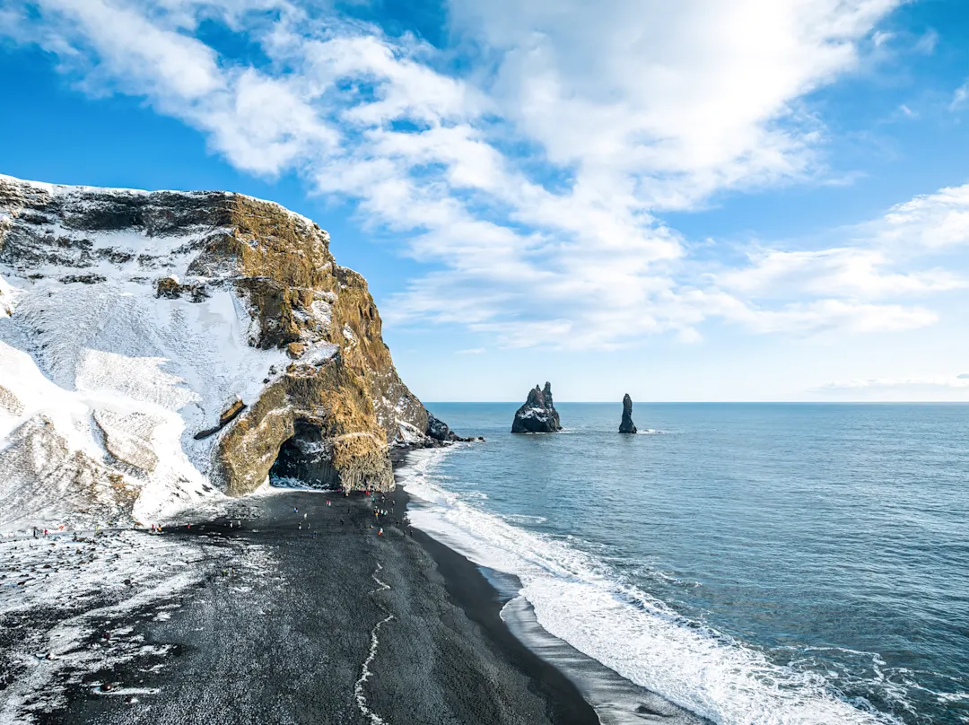 Reynisfjara, Island Schwarzer Sandstrand mit schneebedeckten Klippen und Felsformationen im Meer, Reynisfjara, Island.