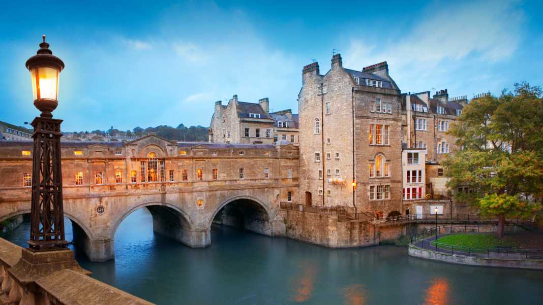 Europe- England, Bath, Pulteney Bridge over a calm river.  Sky approaches evening light, and an old fashioned lamp illuminates the way.