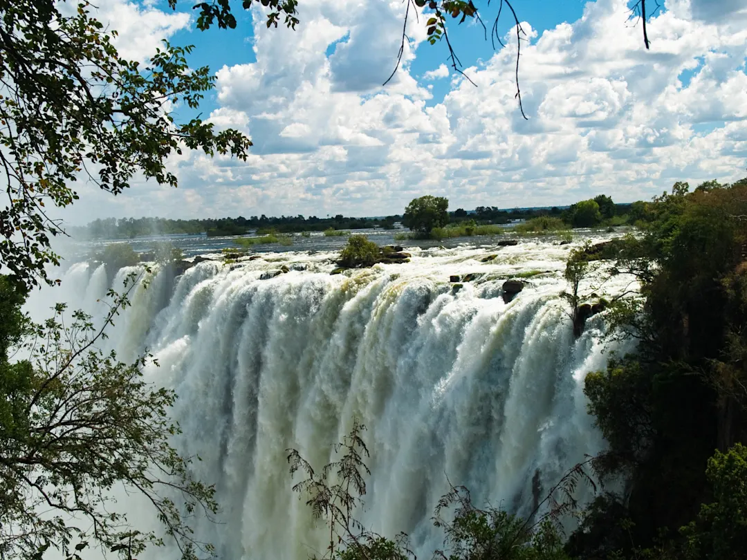 Mächtiger Wasserfall mit weißem Wasser stürzt über eine breite Klippe, umrahmt von grüner Vegetation unter blauem Himmel mit Wolken.