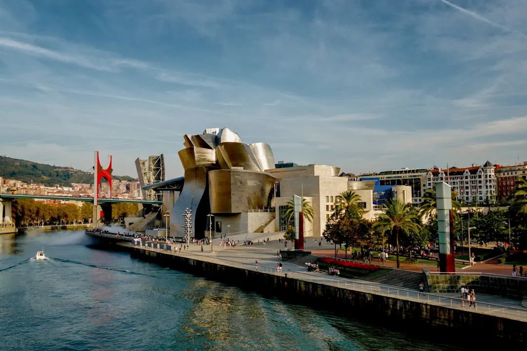 Das Bild zeigt das Guggenheim-Museum Bilbao mit seinem geschwungenen, metallischen Äußeren am Fluss Nervión, blauem Himmel mit Wolken, grünen Hügeln und städtischen Gebäuden im Hintergrund.