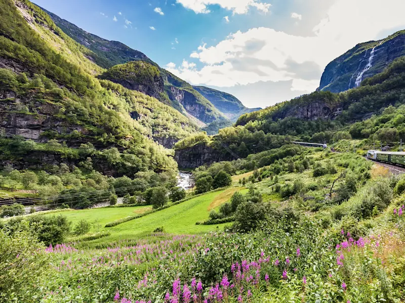 Spektakuläre Zugstrecke durch grünes Tal mit Wasserfall. Flåmsdalen, Vestland, Norwegen.