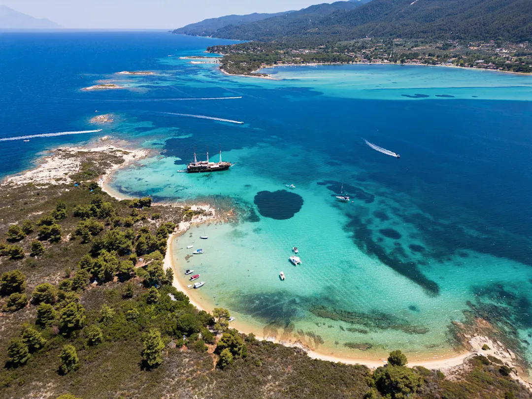 Luftaufnahme einer Küstenbucht mit türkisblauem Wasser, kleinen Booten und einem größeren Schiff, umgeben von bewaldeten Bergen.
