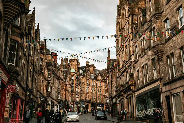 Eine Straße mit Kopfsteinpflaster, gesäumt von historischen Steingebäuden, mit bunten Wimpeln über dem Kopf, parkenden Autos am Straßenrand und Menschen, die unter einem bewölkten Himmel auf den Bürgersteigen spazieren gehen.