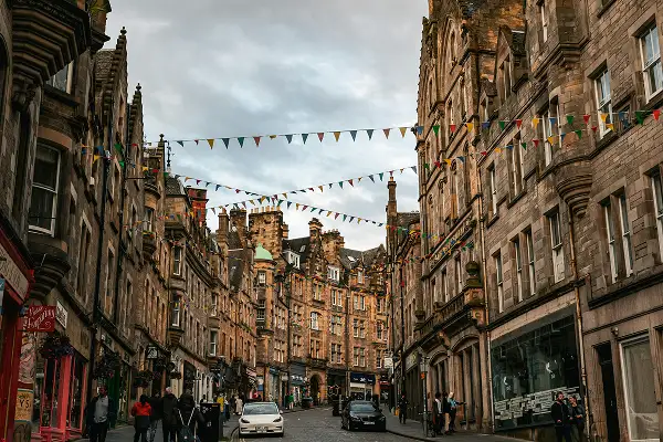 Historic Edinburgh street with stone buildings, colorful bunting strung overhead, and pedestrians walking on cobblestone road.