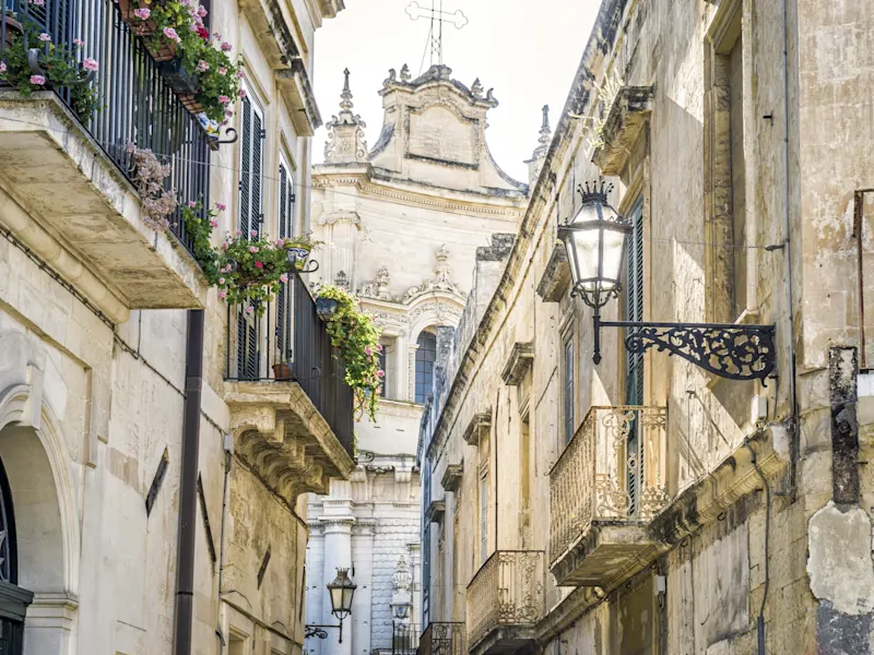 Enge Straße mit Balustraden und Laternen, im Hintergrund die Kirche, Lecce, Apulien, Italien.
