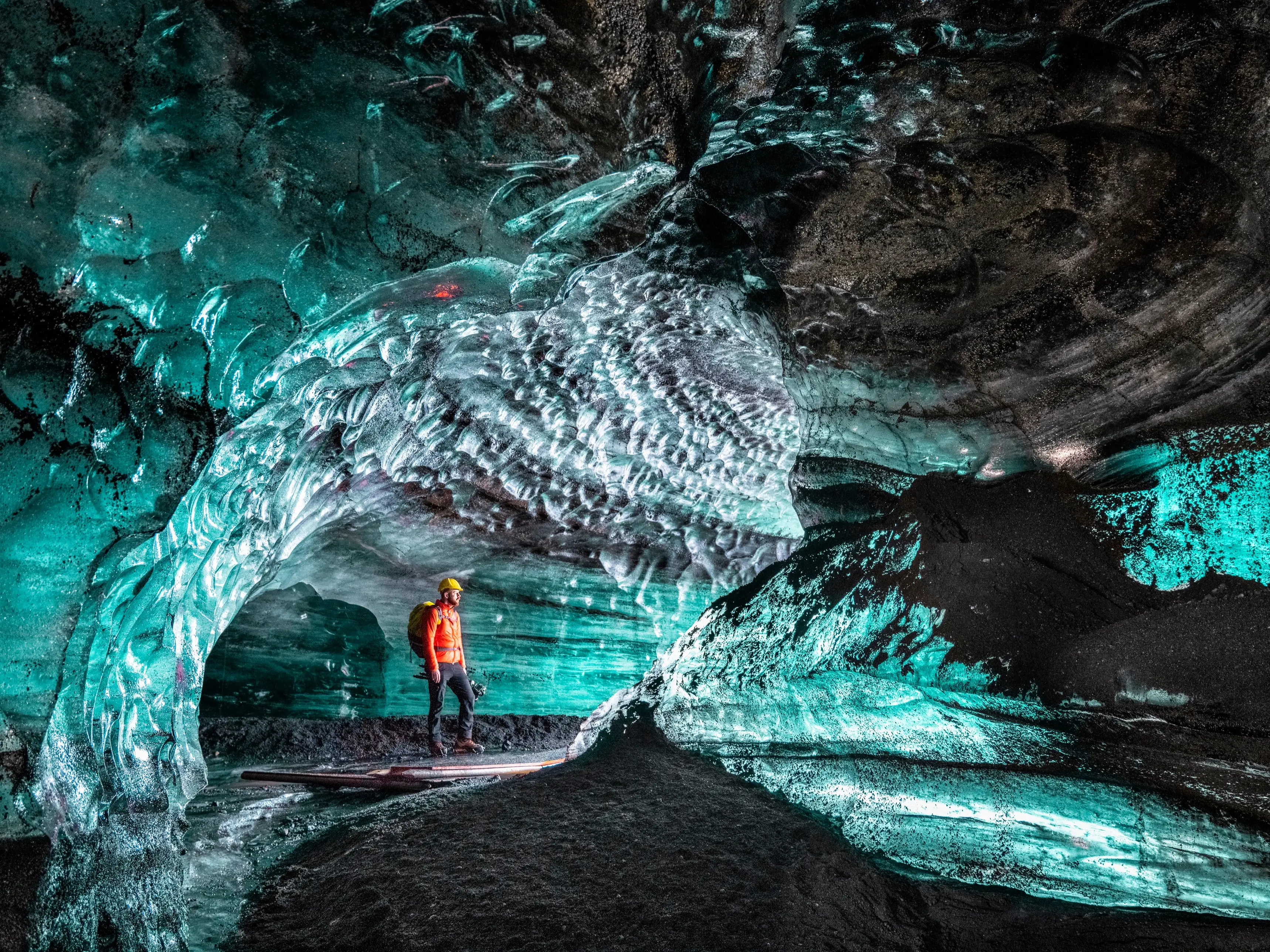 Eine Person in roter Jacke und gelbem Helm steht in einer beleuchteten Eishöhle, umgeben von blau und türkis schimmernden Eisformationen und dunklem felsigem Boden.