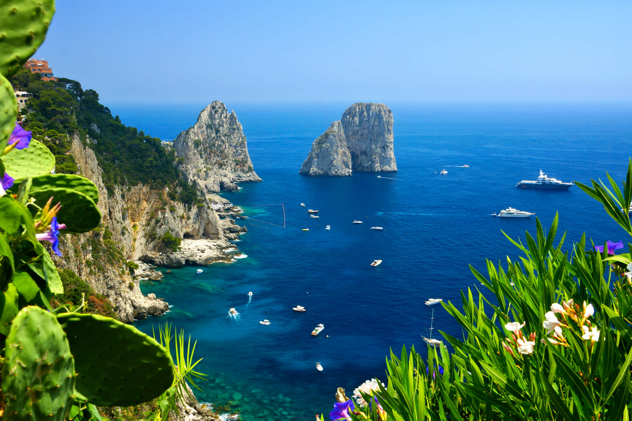 Blick auf die Küste von Capri mit den Faraglioni-Felsen, Blumen und Booten im blauen Meer in Italien
