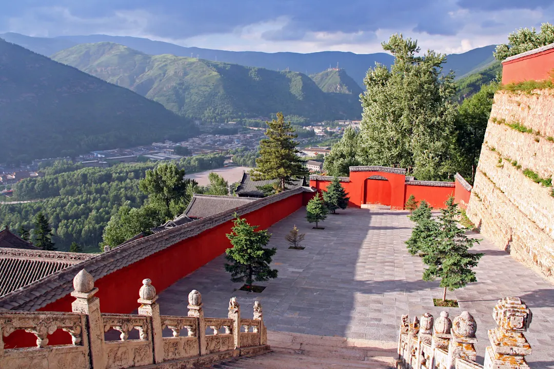 Buddhistischer Nanshan-Tempel in Wutaishan, China. Im Vordergrund Tempelgebäude, im Hintergrund Luftaufnahme der Stadt Wutai Shan und der Berge, die von der untergehenden Sonne beleuchtet werden.


