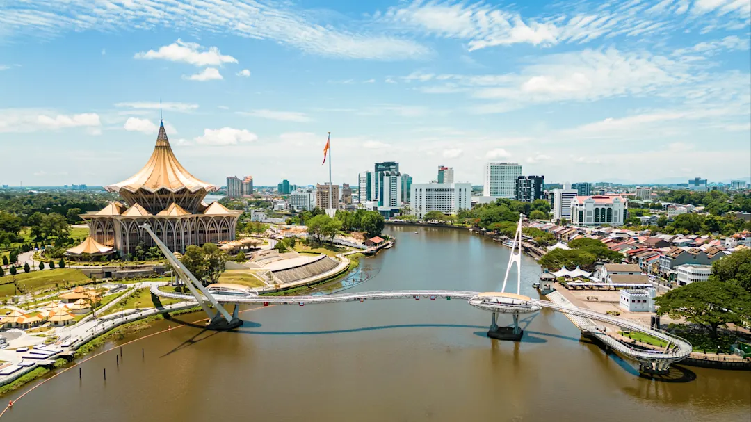 Moderne Skyline mit einer geschwungenen Brücke über den Fluss vor dem Parlament. Kuching, Sarawak, Malaysia.
