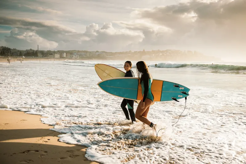 Sydney, New South Wales, Australien Zwei Surferinnen mit Brettern gehen am Bondi Beach bei Sonnenuntergang ins Wasser. Sydney, New South Wales, Australien.