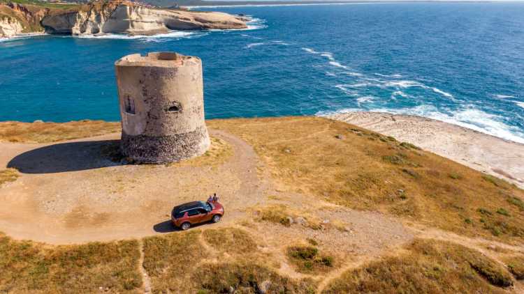 Turm am Rande einer Klippe in Sardinien und zwei Menschen am Auto als Symbolbild für einen Roadtrip durch Italien