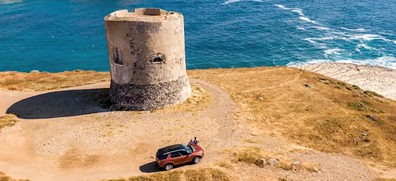 Turm am Rande einer Klippe in Sardinien und zwei Menschen am Auto als Symbolbild für einen Roadtrip durch Italien. Sardinien, Italien