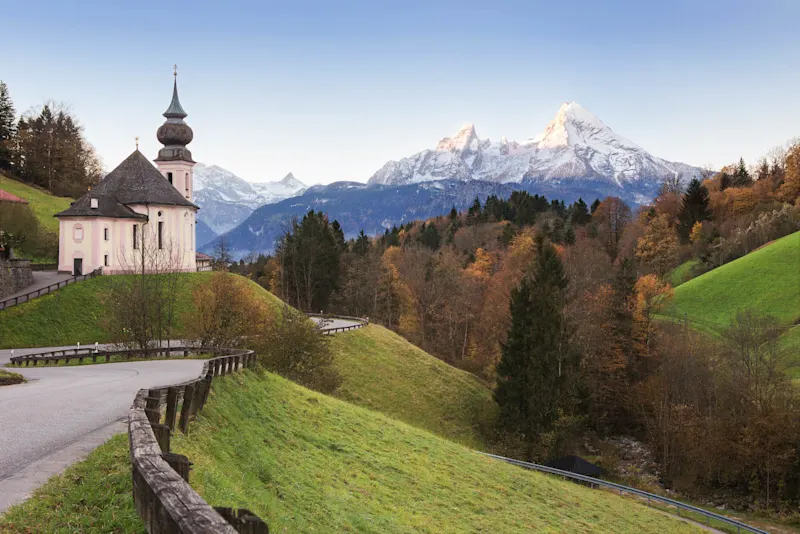 Historic church with onion dome overlooking autumn forest and snow-capped Alpine mountains in Bavaria.