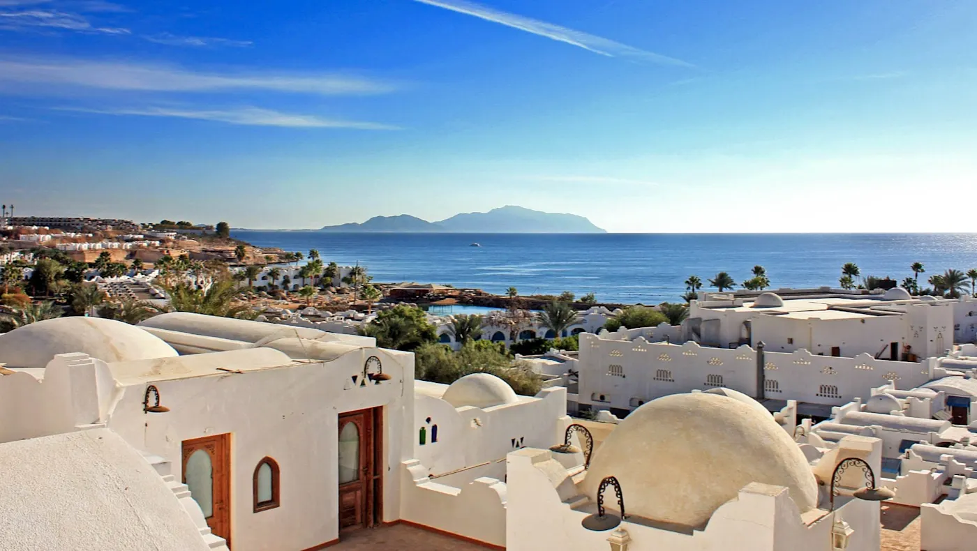 Blick auf das Meer mit Küste und Berg Sinai im Hintergrund, weiße Gebäude mit Kuppeln im Vordergrund unter blauem Himmel, in Sharm El-Sheikh, Ägypten.