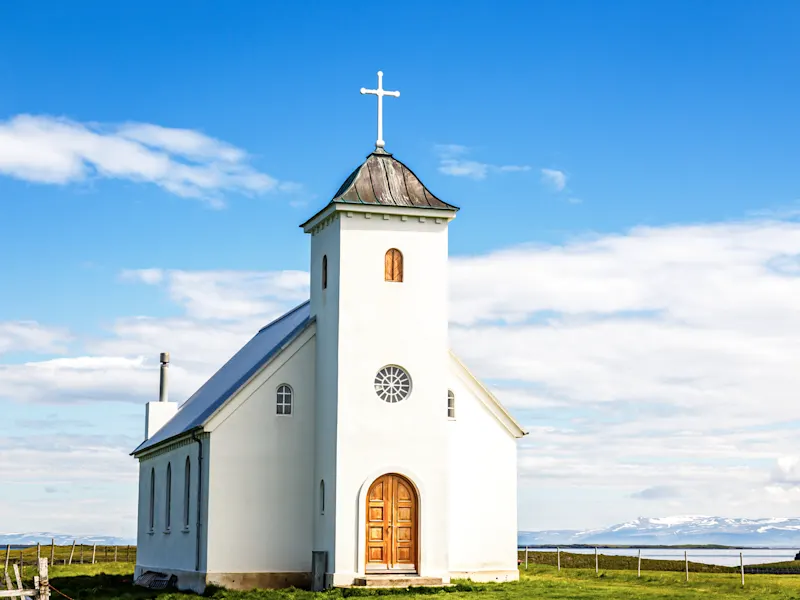 Weiße Kirche auf einer grünen Wiese mit Blick auf Berge und Meer unter blauem Himmel, Westfjorde, Island.