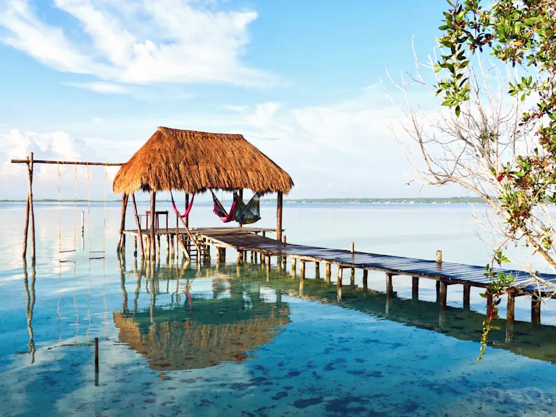 Stimmungsvoller Steg mit Strohdach, Hängematten und Spiegelung im Wasser. Bacalar, Quintana Roo, Mexiko.