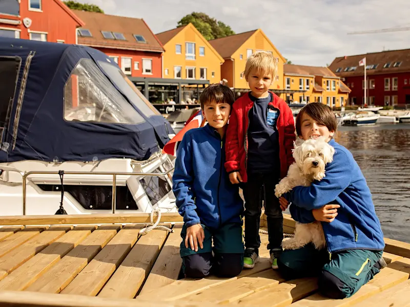 Norwegen, Familienurlaub Kinder und ein kleiner weißer Hund auf einem Steg am Wasser mit bunten Gebäuden im Hintergrund. Norwegen.