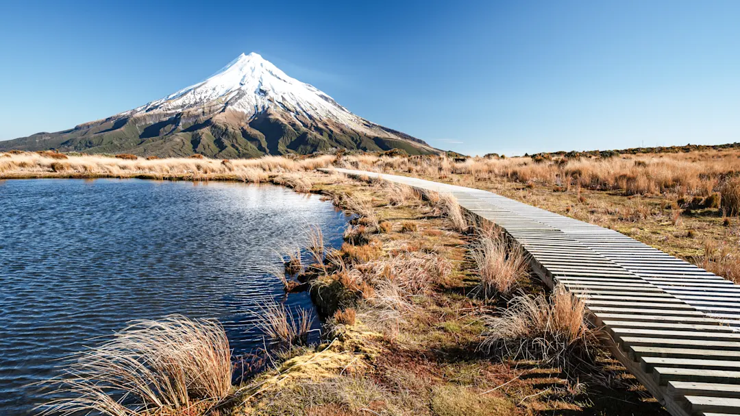 Holzsteg führt entlang eines Sees zu einem schneebedeckten Vulkan unter klarem Himmel. Mount Taranaki, Neuseeland.