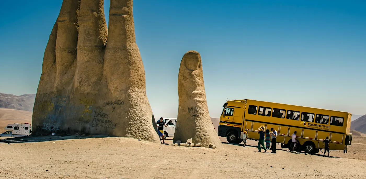 Monumentale Handskulptur in der Wüste mit gelbem Touristenbus daneben, unter blauem Himmel.