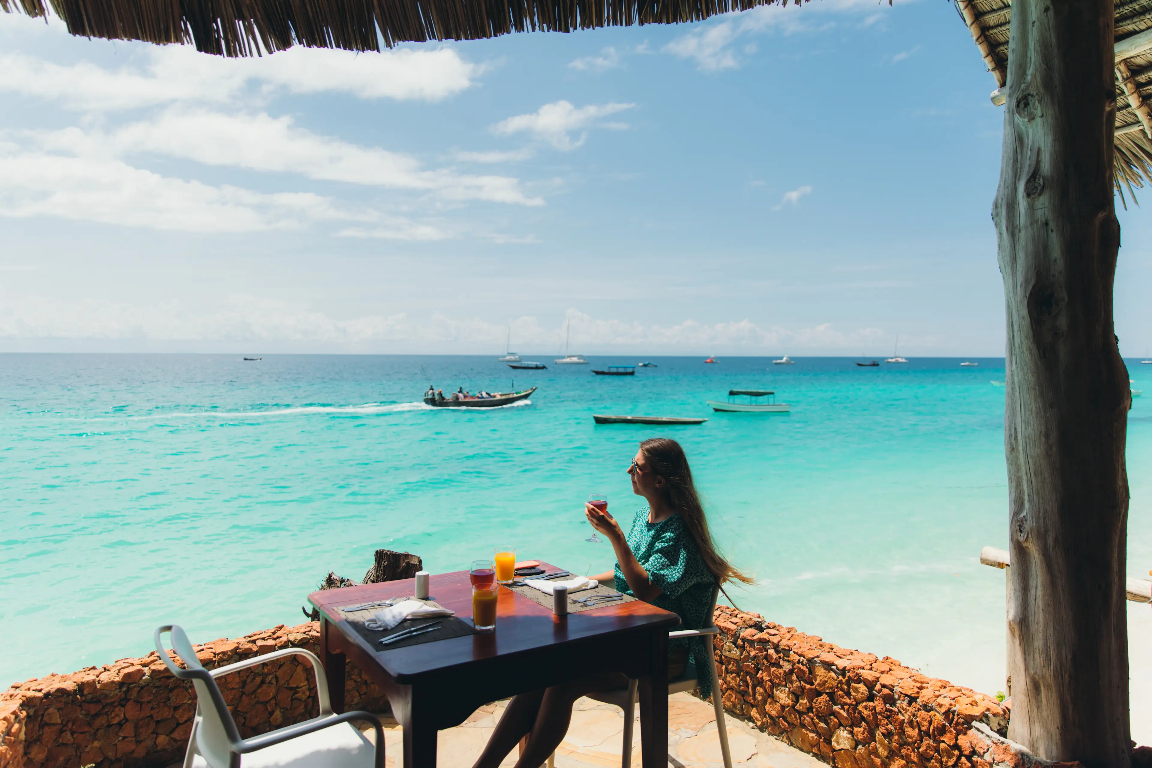 Junge Frau mit langen Haaren im roten Badeanzug genießt Urlaub auf der Insel, blickt von der Restaurantterrasse auf das kristallblaue Meer und die Boote, trinkt Wein und Saft