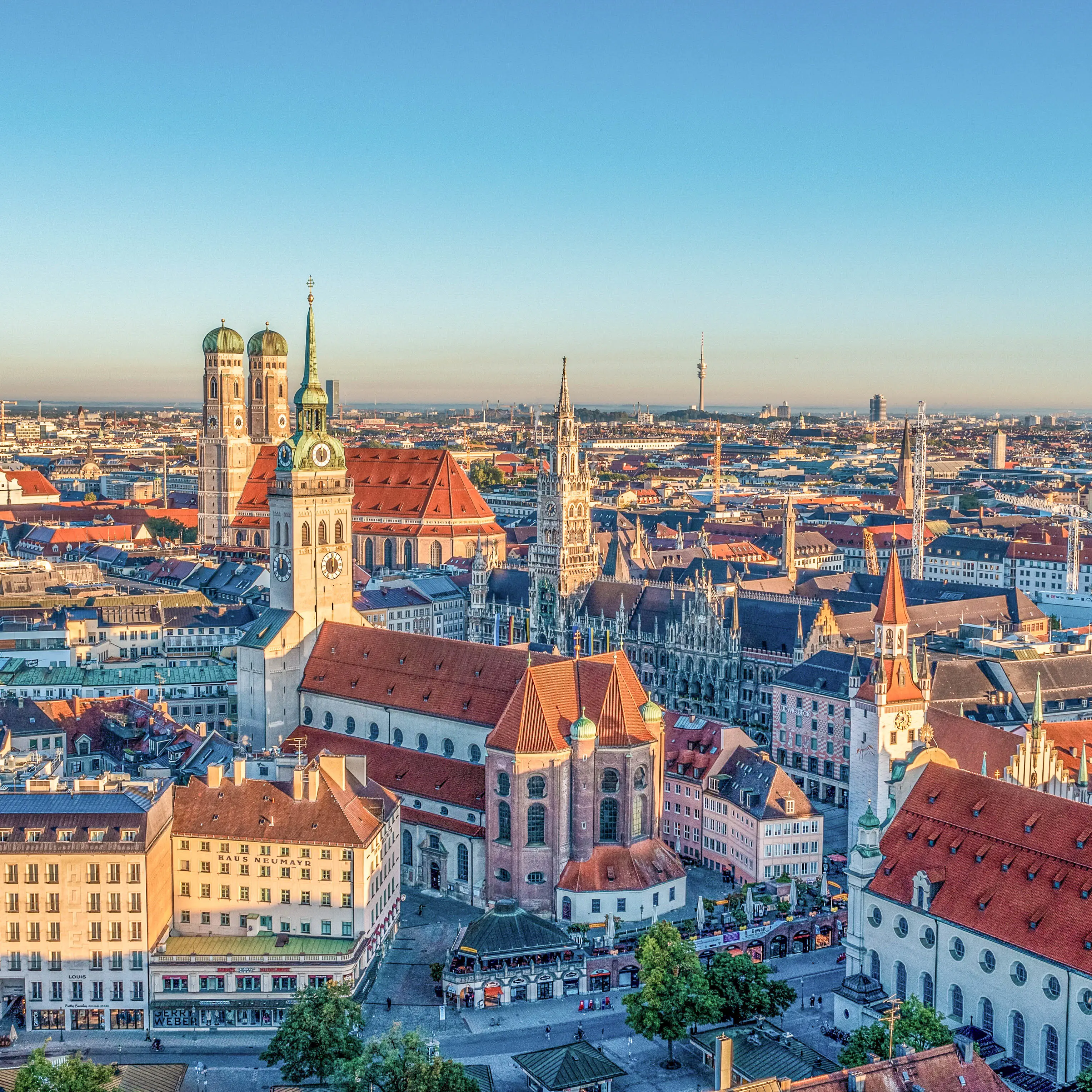 Aerial view of Munich's historic center with Frauenkirche's twin domes and New Town Hall against clear blue sky at sunset.