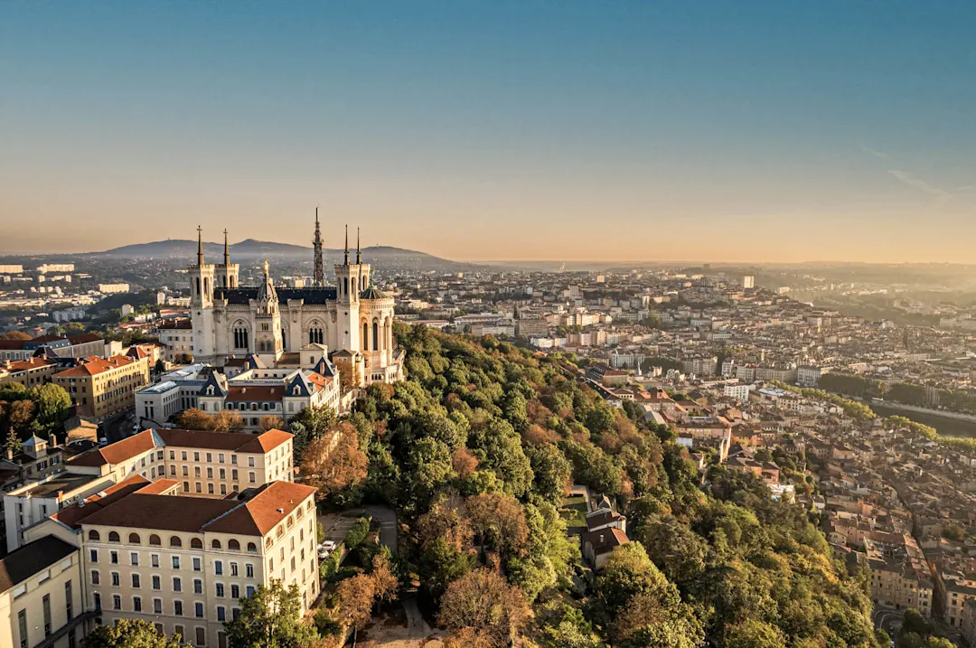 Luftaufnahme von Lyon mit der Basilika Notre-Dame de Fourvière bei Sonnenuntergang, Stadtpanorama und Hügeln im Hintergrund.