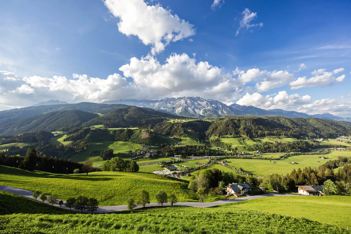 Scenic Alpine valley with lush green meadows, scattered houses, forested hills, and snow-capped mountains under a blue sky with clouds.