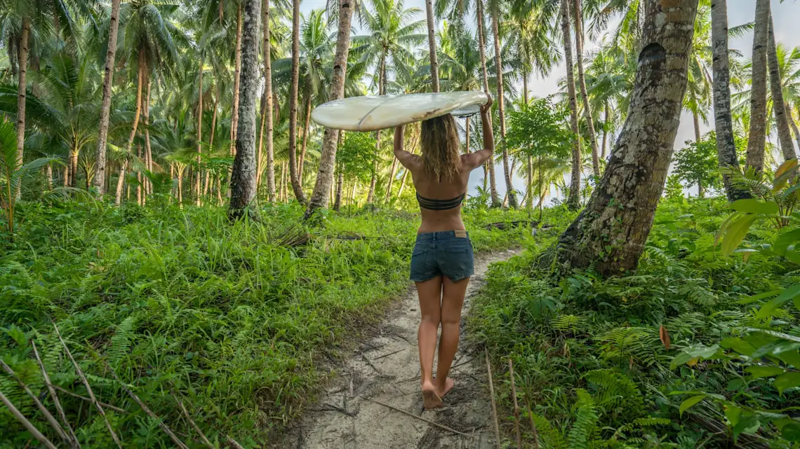 Eine Frau trägt ihr Surfbrett über dem Kopf und läuft durch Palmen Richtung Strand, Philippinen.
