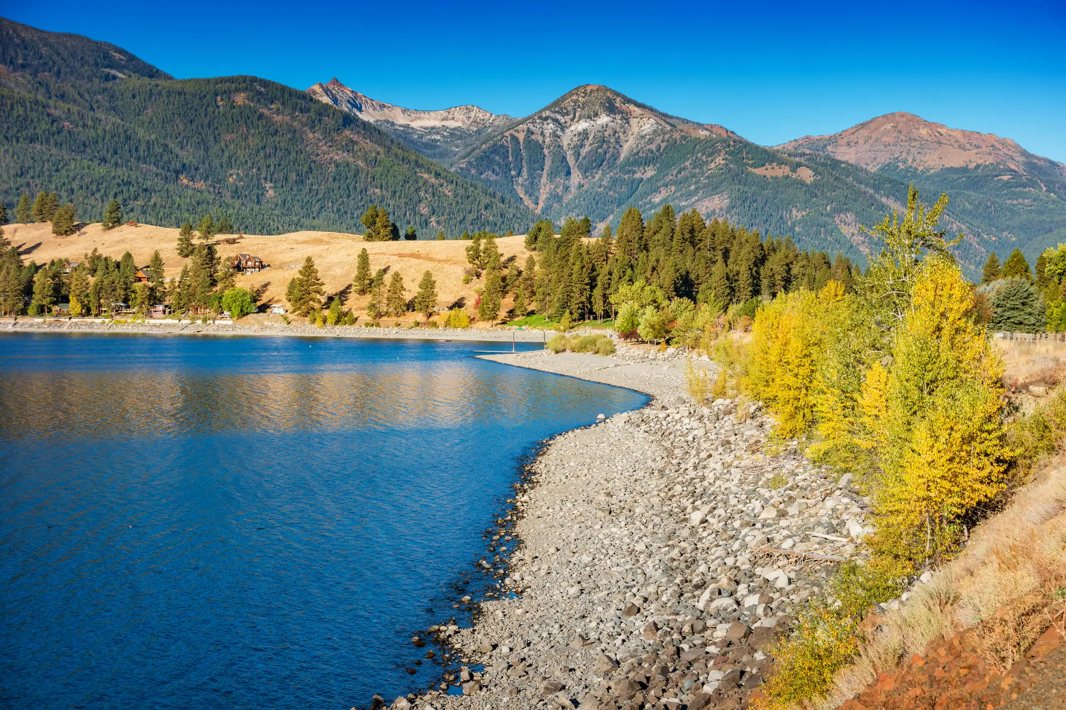 Blauer Bergsee mit steinigem Ufer, grünen Bäumen und goldenen Wiesen vor Bergkulisse, Joseph, Oregon, USA. 