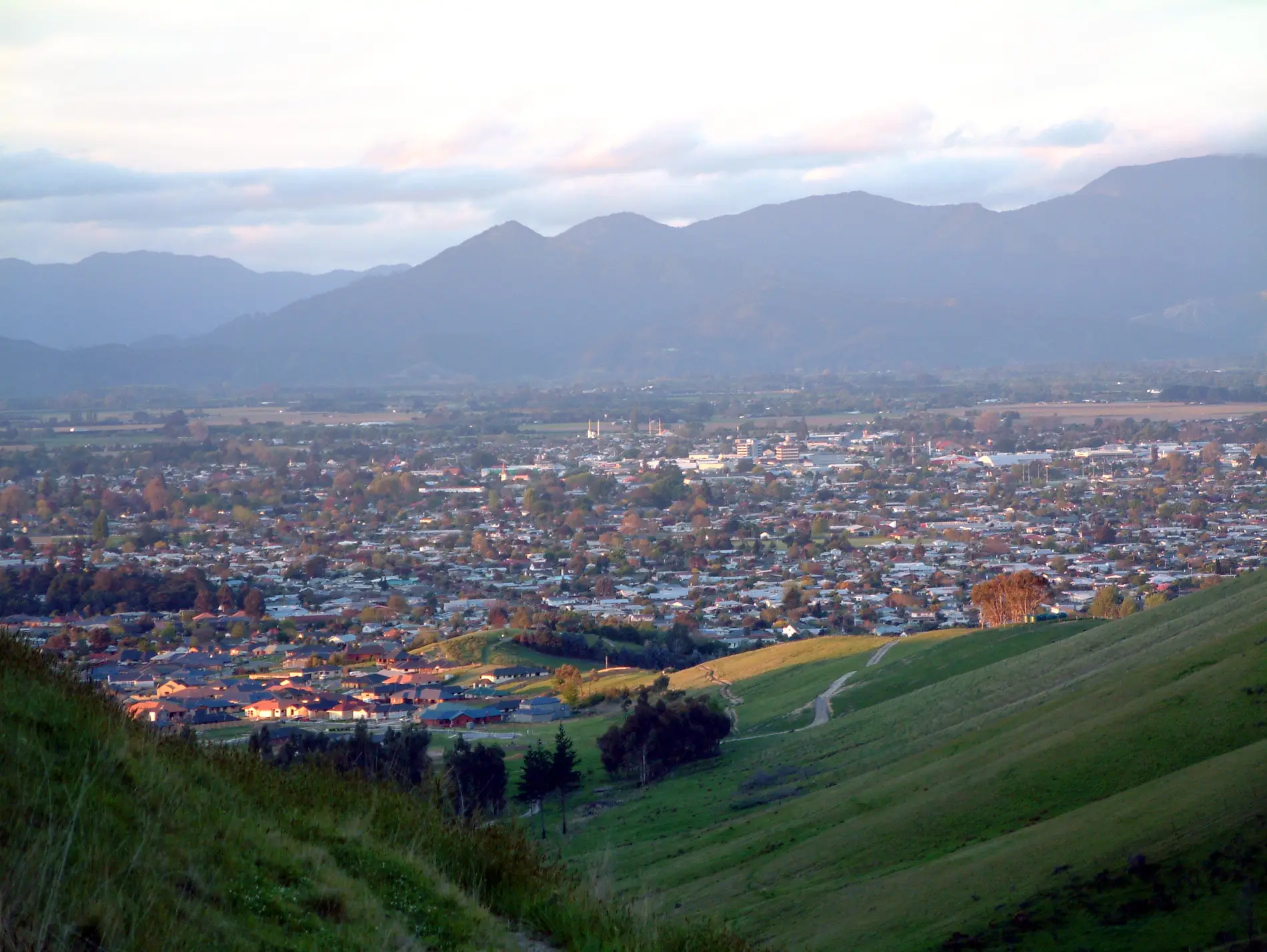 Eine Stadt schmiegt sich in ein weites Tal, umgeben von grünen Hügeln und fernen Bergen unter einem teilweise bewölkten Himmel, wobei das sanfte Sonnenlicht Teile der Landschaft beleuchtet.