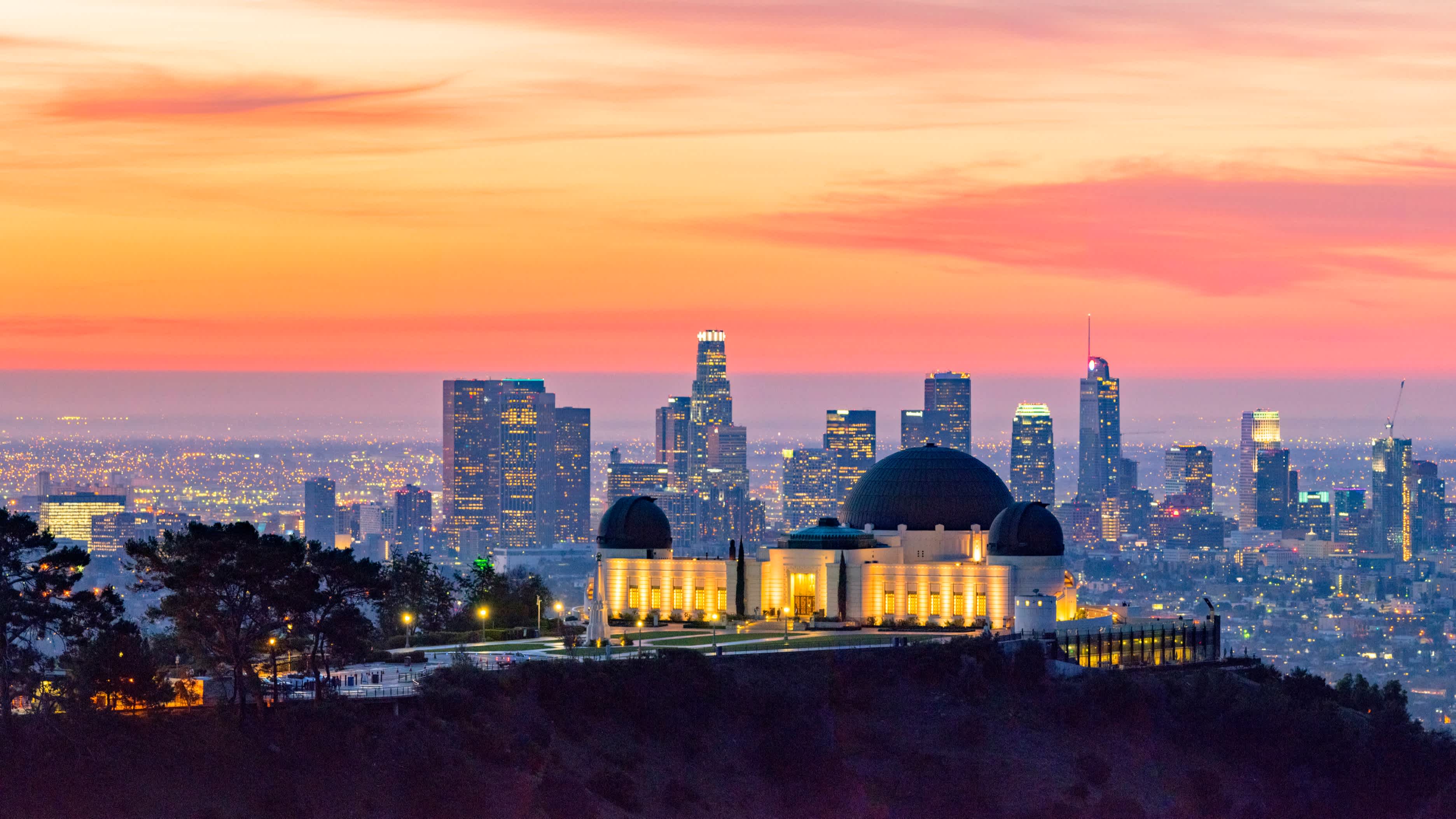 Vue aérienne sur le célèbre observatoire de Griffith Park, un lieu célèbre à visiter pendant votre voyage à Los Angeles, au coucher du soleil avec la skyline de Los Angeles en arrière-plan.