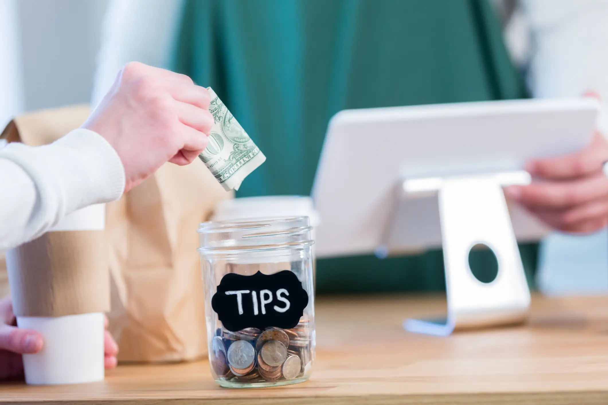 Ein unkenntlicher Coffee-Shop-Kunde steht gegenüber einem nicht erkennbaren Barista an der Kasse und streckt die Hand aus, um eine Papierrechnung in das Trinkgeldglas zu legen.