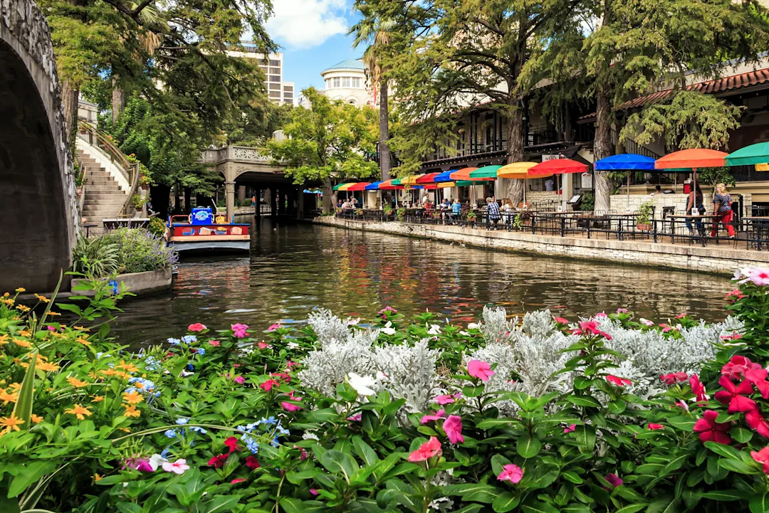 Angelegte Beete mit Blumen an einem Kanal in San Antonio mit Tischen und bunten Sonnenschirmen entlang des Ufers