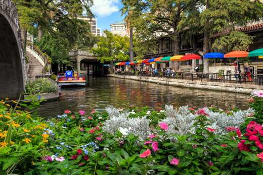Angelegte Beete mit Blumen an einem Kanal in San Antonio mit Tischen und bunten Sonnenschirmen entlang des Ufers