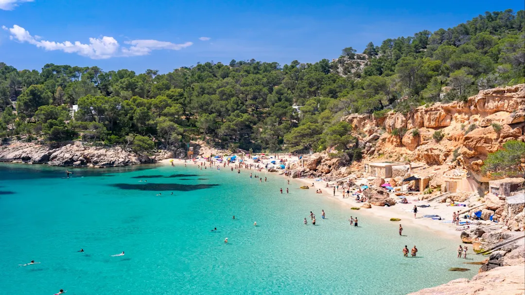Weitwinkelblick auf Cala Saladeta, nur wenige Schritte von Cala Salada entfernt, an der Westküste Ibizas, bekannt für sein extrem transparentes Wasser, den perlmuttfarbenen Sand und eine malerische Gruppe von Fischerhütten (lokal bekannt als Casitas Varadero). Badegäste mit verschiedenen Aktivitäten, dem hellen Licht eines Sommermittags, malerischen Wolken, bunten Klippen, üppiger Vegetation.