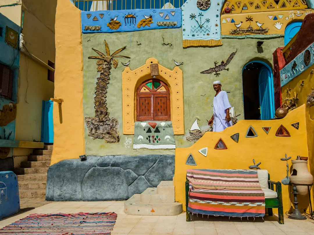 Assuan, Ägypten Ein farbenfrohes nubisches Haus mit traditionellen Mustern, bunten Stoffen und Lehnstuhl, ein Mann in traditioneller Kleidung davor, in Assuan, Ägypten.