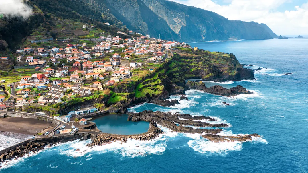 Bunte Häuser auf terrassierten Klippen am Meer mit Bergen im Hintergrund, São Vicente, Madeira, Portugal.

