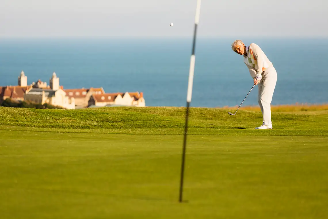 Joueur de golf, Angleterre Golfeur en tenue blanche jouant sur un parcours côtier avec vue sur la mer et un bâtiment historique en arrière-plan.