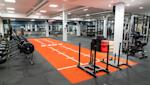 Modernt gym med orange träningsyta, roddmaskiner, viktställ och hantlar under rosa belysning i industriell lokal.