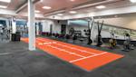 Modernt gym med orange löparbana, roddmaskiner och träningsutrustning, grått golv och synliga ventilationskanaler.