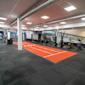 Modernt gym med orange löparbana, roddmaskiner och träningsutrustning, grått golv och synliga ventilationskanaler.